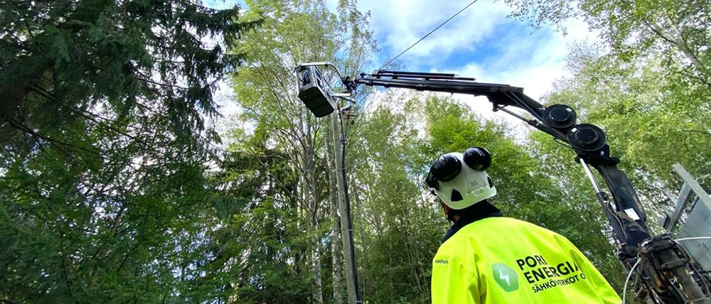 Ilmajohtoa korjataan, kuvassa Pori Energia Sähköverkkojen työntekijä selin kameraan seuraa nostokorissa työskentelevän työntekoa metsässä.